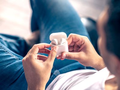 A man in jeans and a white T-shirt lying down and opening an Apple AirPods charging case with two AirPods earphones inside.