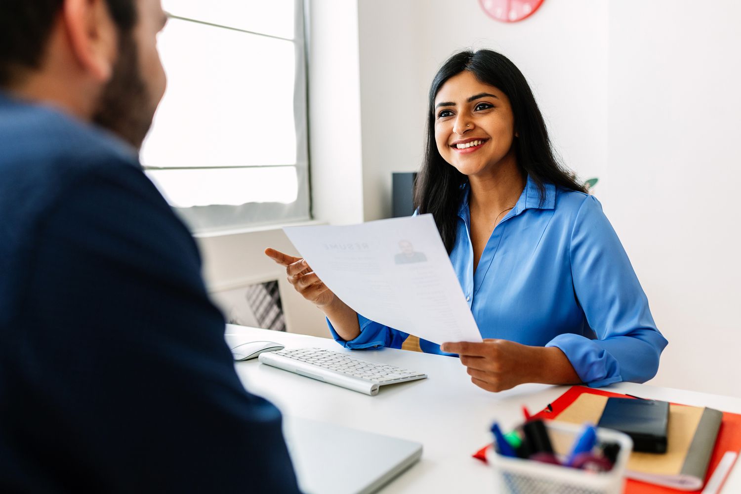 A hiring professional holding a resume and speaking with a candidate in an office setting. 